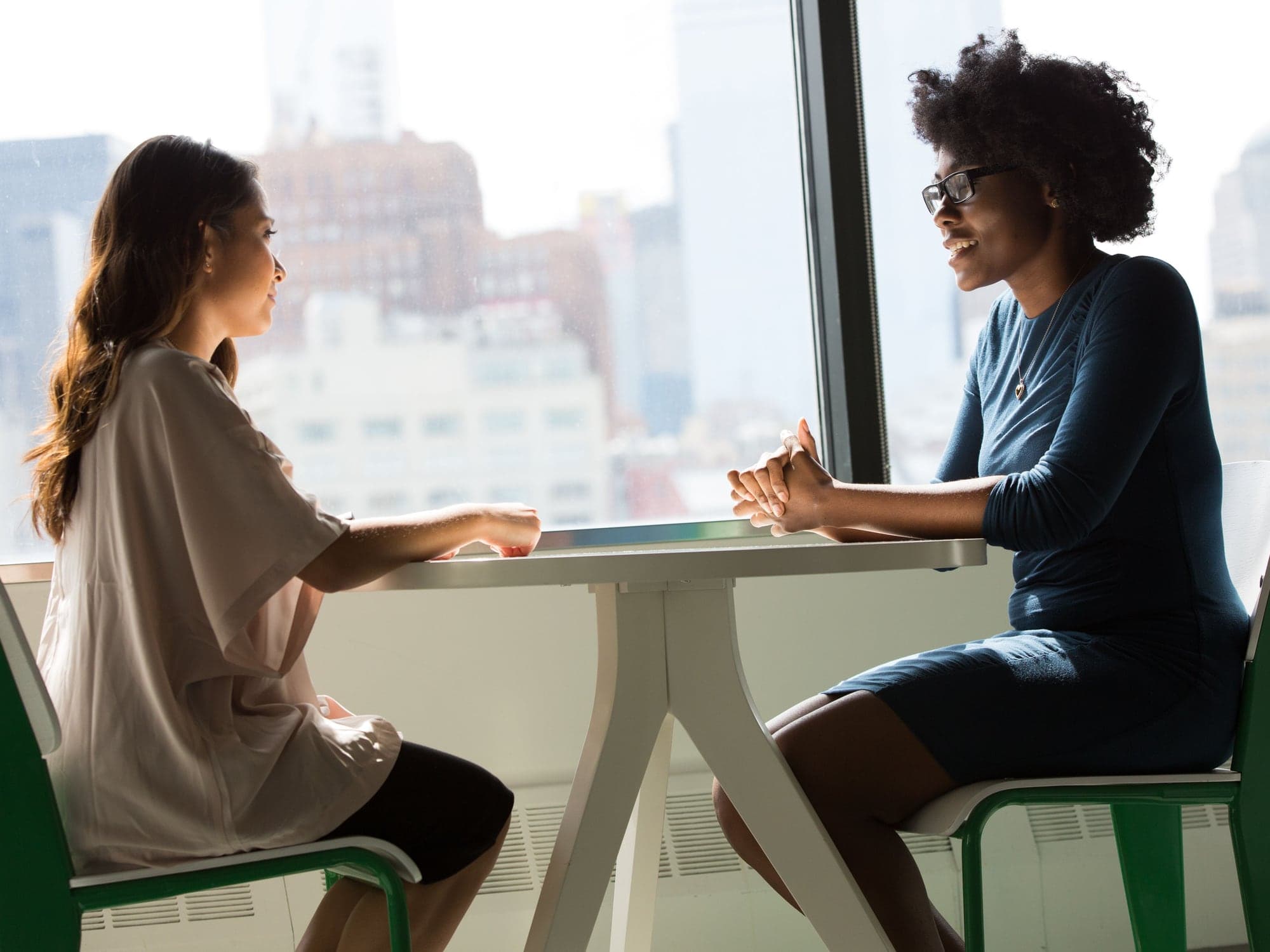 Two adults talking across a white table near a window