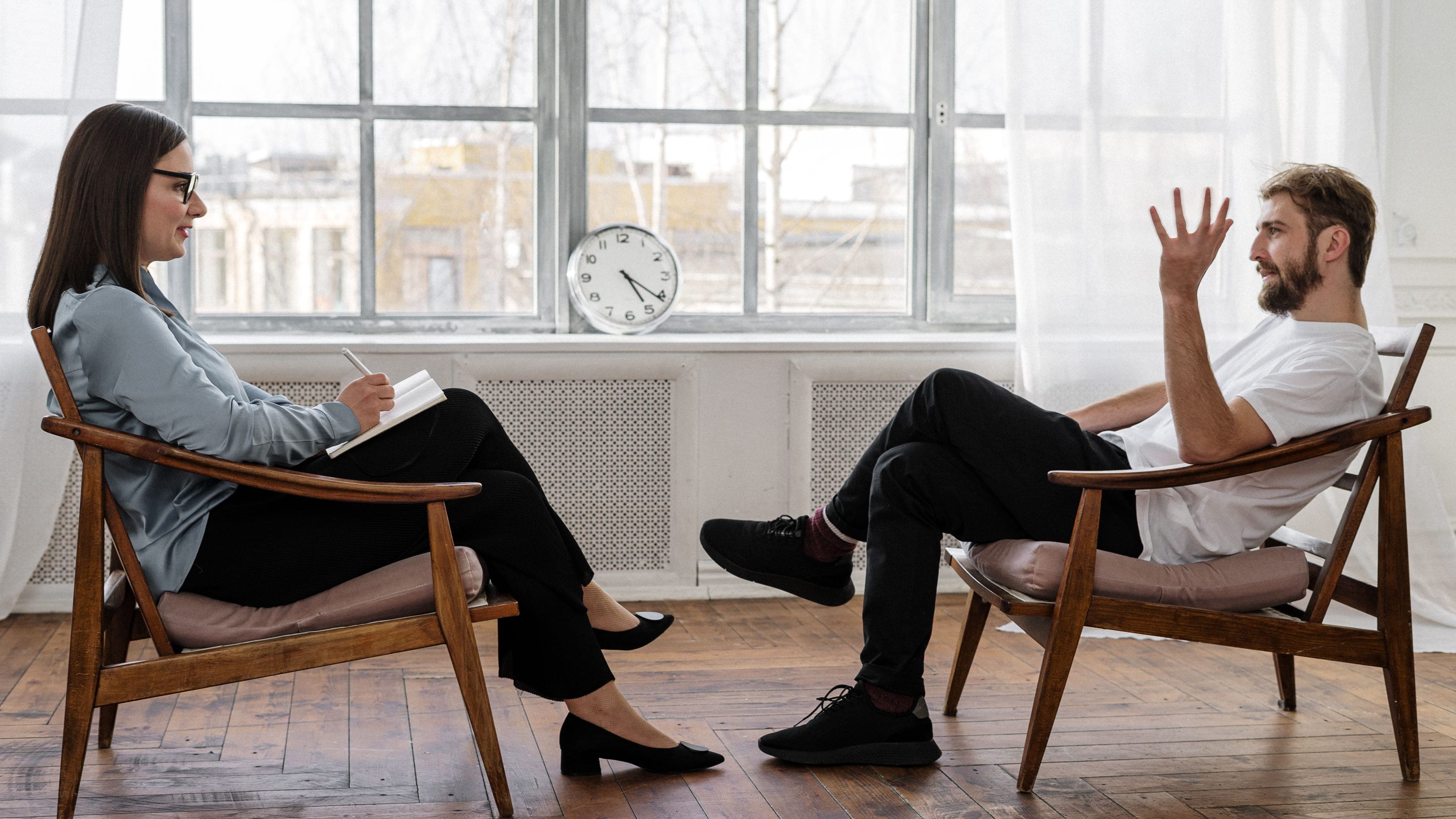 Two people in conversation across chairs near a window during a consultation