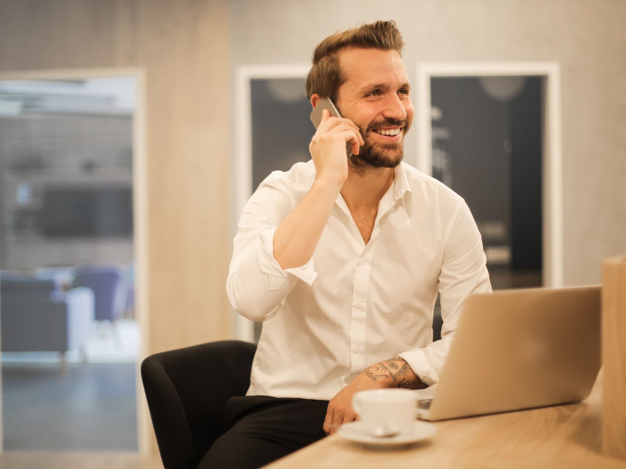 A man in a white shirt speaking on a phone at a desk beside a laptop