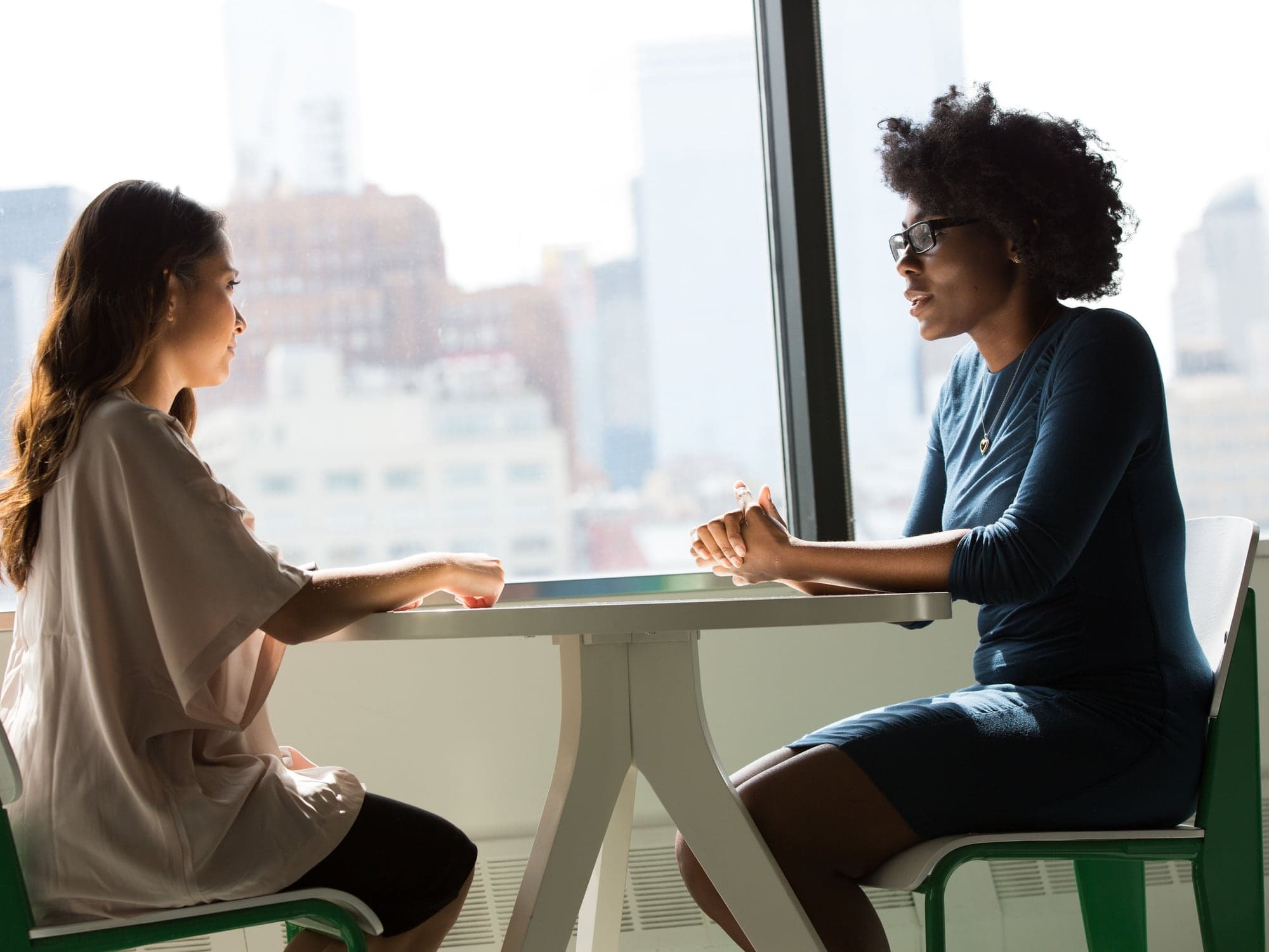 Two adults talking across a white table near a window